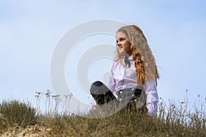 A girl in a white shirt with a bow tie sits on the ground in a field against the background of the sky