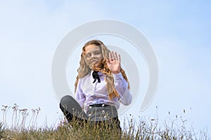 A girl in a white shirt with a bow tie sits on the ground in a field against the background of the sky and waves a pen to the