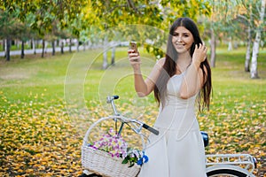 Girl in a white skirt and a vest with a smartphone in her hands stands next to a bicycle and a basket of flowers on yellow leaves