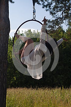 A girl in a white stage costume performs exercises in an aerial ring in the open air, against the background of the forest and the