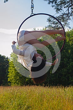 A girl in a white stage costume performs exercises in an aerial ring in the open air, against the background of the forest and the