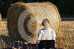 Girl in a white sweater on the background of a haystack and green forest in a field works at a laptop