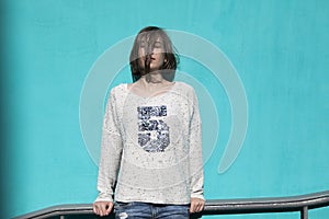 Girl in a white sweater dreamily closes her eyes from the bright sun opposite the blue wall in the underpass