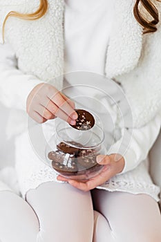 A girl in a white sweater, skirt and tights holds a glass jar with chocolate chip cookies decorated with stars.