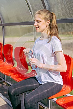 A girl in a white T-shirt and black leggings sits on red chairs for fans and holds a water bottle in her hands