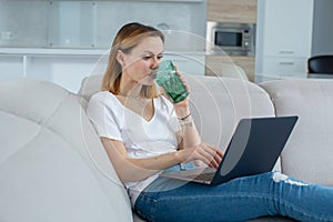 A girl in a white T-shirt drinks water from a glass, works behind a laptop