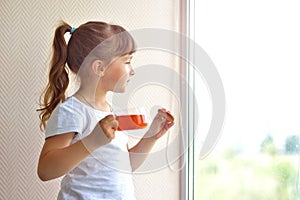 A girl in a white T-shirt holds a cup of tea and a liver in the form of a red heart standing by the window, watching. delicious