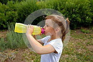 A girl in a white T-shirt in the summer on the street drinks water from a yellow plastic bottle of ecological