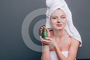 Girl with a white towel on her head with a nutritious green mask on her face and an avocado in her hands on a green background wit