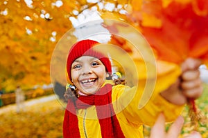 A girl with a wide open smile is playing with red and yellow leaves in an autumn park.