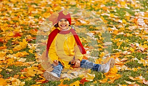A girl with a wide smile is sitting on a carpet of red and yellow leaves in an autumn park.