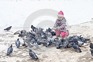 A girl in winter clothes sat down and feeds pigeons with crumbs. saving the pitz from starvation in winter