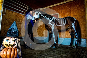 A girl in a witch costume holds a horse by the bridle in a corral on a farm, a skeleton is painted on a horse in white paint