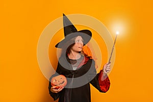 Girl in wizard student costume with luminous magic wand and Halloween pumpkin looking at light on yellow background.