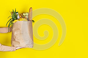 Girl or woman holds a paper bag filled with groceries such as fruits, vegetables, milk, yogurt, eggs isolated on yellow