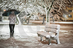 Girl or woman walking on the strees with umbrella in snowstorm, bench with snow in the city, night photography