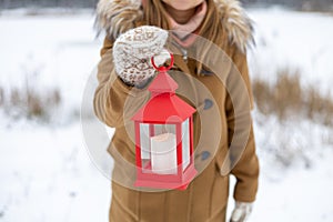 A girl in woolen mittens holds a red lantern in her hands with a candle in the winter forest