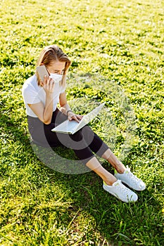 Girl working at computer and talk by phine sitting on grass in park. Freelance, distance education concept.