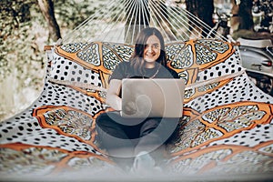 Girl working on laptop while lying on hammock in sunny summer park. freelancer woman hipster with laptop relaxing and smiling.