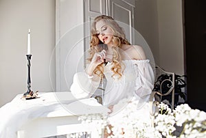 Girl writes a letter to her beloved man, sitting at home at table in a white light dress, purity and innocence. Curly blonde