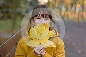 girl in yellow coat sitting on the bench. The girl holds an armful of leaves