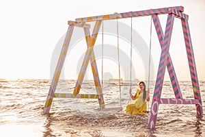 A girl in a yellow dress rides on a swing in the sea among splashes and waves.