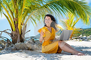 Girl in a yellow dress on a tropical sandy beach works on a laptop and drinks fresh mango. Remote work, successful freelance.