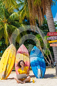 Girl in a yellow dress on a tropical sandy beach works on a laptop near kayaks and drinks fresh mango. Remote work, successful