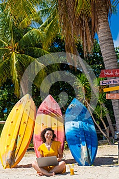 Girl in a yellow dress on a tropical sandy beach works on a laptop near kayaks and drinks fresh mango. Remote work, successful