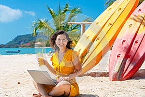 Girl in a yellow dress on a tropical sandy beach works on a laptop near kayaks and drinks fresh mango. Remote work, successful