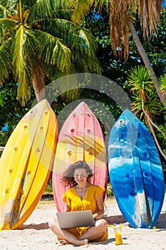 Girl in a yellow dress on a tropical sandy beach works on a laptop near kayaks and drinks fresh mango. Remote work, successful