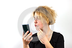 A girl with yellow hair talks on a video link on a smartphone, and examines the pimples on her face, a thoughtful