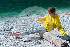 A girl in a yellow hoodie sits on a log by the sea and balances while keeping her legs hanging