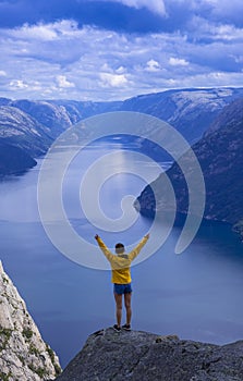 Girl in yellow jacket with arms raised looking at Lysefjord fjord from Preikestolen.