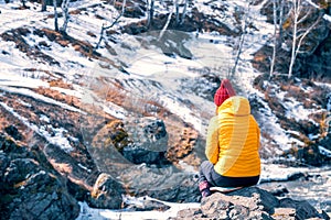 Girl in a yellow jacket and a red knitted hat in nature sits on a stone. rest in the highlands in spring. reflections on life in