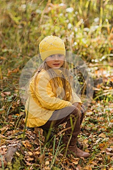 Girl in a yellow jacket and a yellow hat sitting on a tree stump in the autumn forest a sunny day
