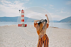 A girl in a yellow-orange suit on the beach against the background of the sea, mountains and a lighthouse. Fashion woman in