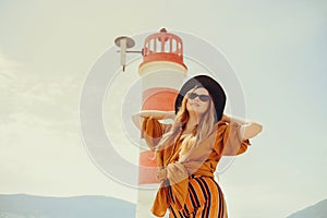 A girl in a yellow-orange suit on the beach against the background of the sea, mountains and a lighthouse. Fashionable woman in