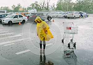 Girl in yellow raincoat stand alone under rain in parking lot. Look down on wet asphalt. Many cars behind. Empty