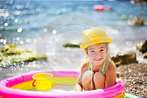 Girl in yellow straw hat plays in outdoor near sea, in water with a bucket in an inflatable pool on the beach. Indelible products