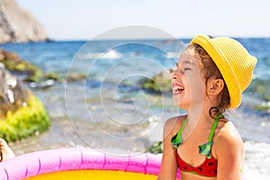 Girl in yellow straw hat plays in outdoor near sea, in water with a bucket in an inflatable pool on the beach. Indelible products