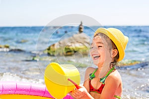 Girl in yellow straw hat plays in outdoor near sea, in water with a bucket in an inflatable pool on the beach. Indelible products