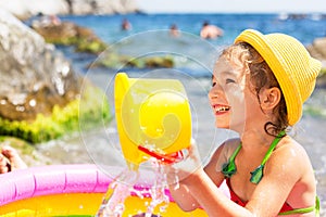 Girl in yellow straw hat plays in outdoor near sea, in water with a bucket in an inflatable pool on the beach. Indelible products