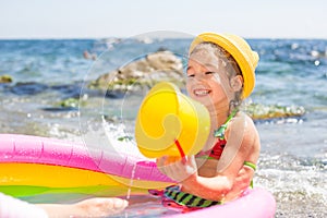 Girl in yellow straw hat plays with the wind, water and a water dispenser in an inflatable pool on the beach. Indelible products