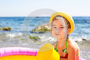 Girl in yellow straw hat sitting in an inflatable pool by the sea with a serious and dissatisfied look. Indelible products to