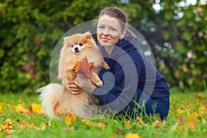 Girl or young woman holding Pomeranian spitz and bouquet of fallen leaves in the park in autumn