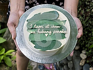 Girlfriend handing over a small cake with green icing and a heartwarming message. Tagalog words translated as 1 year and forever. photo