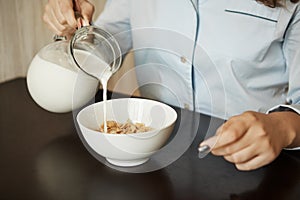 Girlfriend preparing simple breakfast in morning. Cropped shot of woman in nightwear pouring milk in bowl with cereals