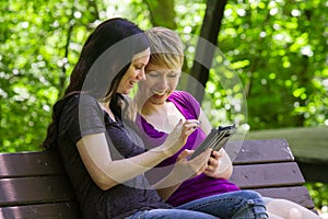 Girlfriends sharing an etablet on park bench, horizontal