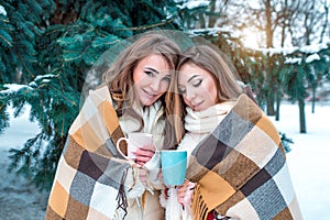 Girlfriends winter in the forest on background of snow and fir trees. Warmed by a rug, in the hands of a mug with a hot